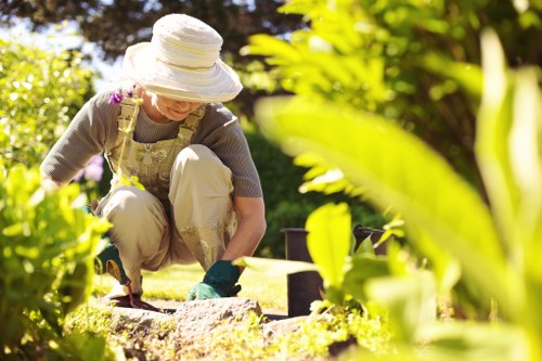 A woman wearing a wide-brimmed sun hat, gloves, and casual gardening attire is kneeling on a neatly maintained grassy garden area in Waterloo, tending to a flower bed bordered by small, dark edging stones. Behind her, there are lush green bushes and trees, with sunlight filtering through the leaves, indicating a bright, clear day. The garden features a well-kept lawn, wooden planters, and a variety of plants, suggesting regular outdoor maintenance. The scene illustrates care in outdoor space management typical of professional gardening services in the Waterloo area, with a focus on healthy plant growth, tidy presentation, and natural floral and foliage tones, fitting seamlessly into the context of local landscaping and garden upkeep provided by Gardening Services Waterloo.