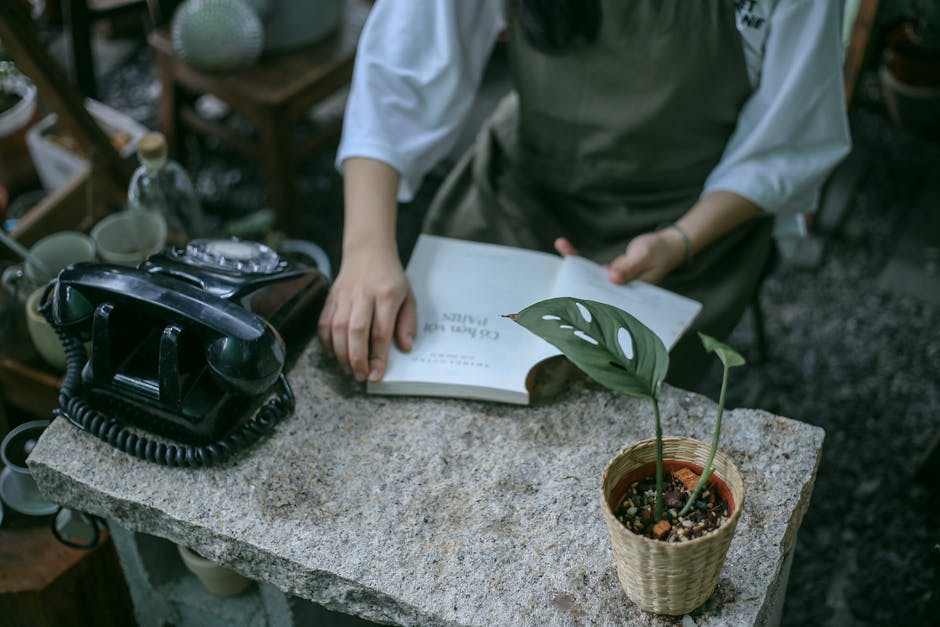 A person wearing a white long-sleeve shirt and green apron is seated at a rustic stone garden table. The table features an vintage black rotary telephone, an open book, and a small potted plant with g