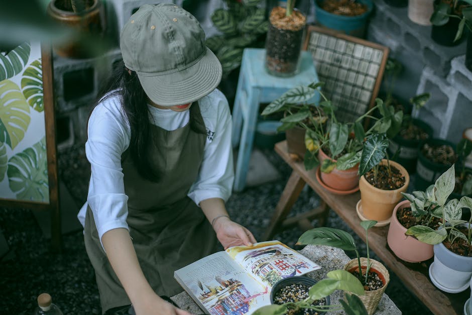 A person wearing a white long-sleeve shirt and green apron is seated at a rustic stone garden table. The table features an vintage black rotary telephone, an open book, and a small potted plant with g