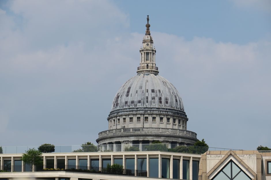 A detailed, long-range view of the historic dome of St. Paul's Cathedral, situated behind a modern rooftop terrace in central London. The foreground features a landscaped garden with well-maintained g